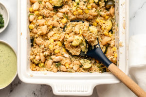 Lemon Dill Quinoa Casserole in white casserole dish with a wooden spoon scooping out the casserole. Small dished of fresh dill and green goddess dressing are off to the side.