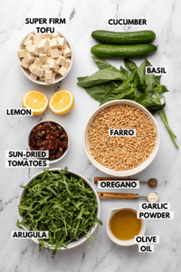 Ingredients for Mediterranean Farro Salad in small white bowls on marble countertop. Clockwise text labels read cucumber, basil, farro, oregano, garlic powder, olive oil, arugula, sun-dried tomatoes, lemon, and super-firm tofu