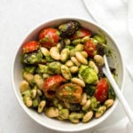 White bean pesto salad with tomatoes and cucumber in a small white bowl, with a small white ceramic spoon. A white linen is off to the right