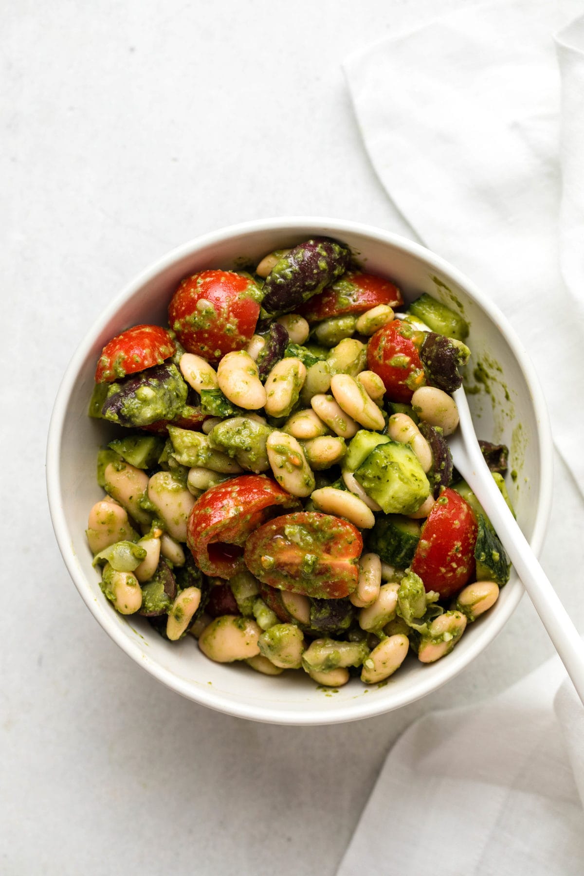 White bean pesto salad with tomatoes and cucumber in a small white bowl, with a small white ceramic spoon. A white linen is off to the right