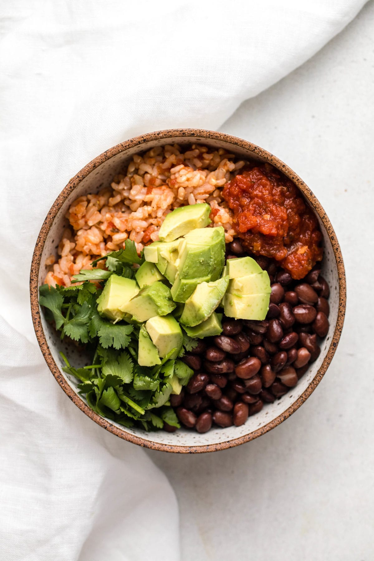 Vegan Burrito Bowl with black beans, salsa, rice, cilantro, and avocado in small speckled bowl on white background