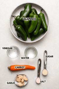 Ingredients for pickled jalapenos arranged on light background. Clockwise text labels read jalapeños, sugar, salt, garlic, carrot, vinegar, and water