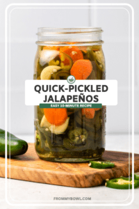 Large glass jar of pickled jalapenos with sliced carrots and garlic in brine. The glass jar is on a small wood cutting board on a white countertop, with white tile in the background