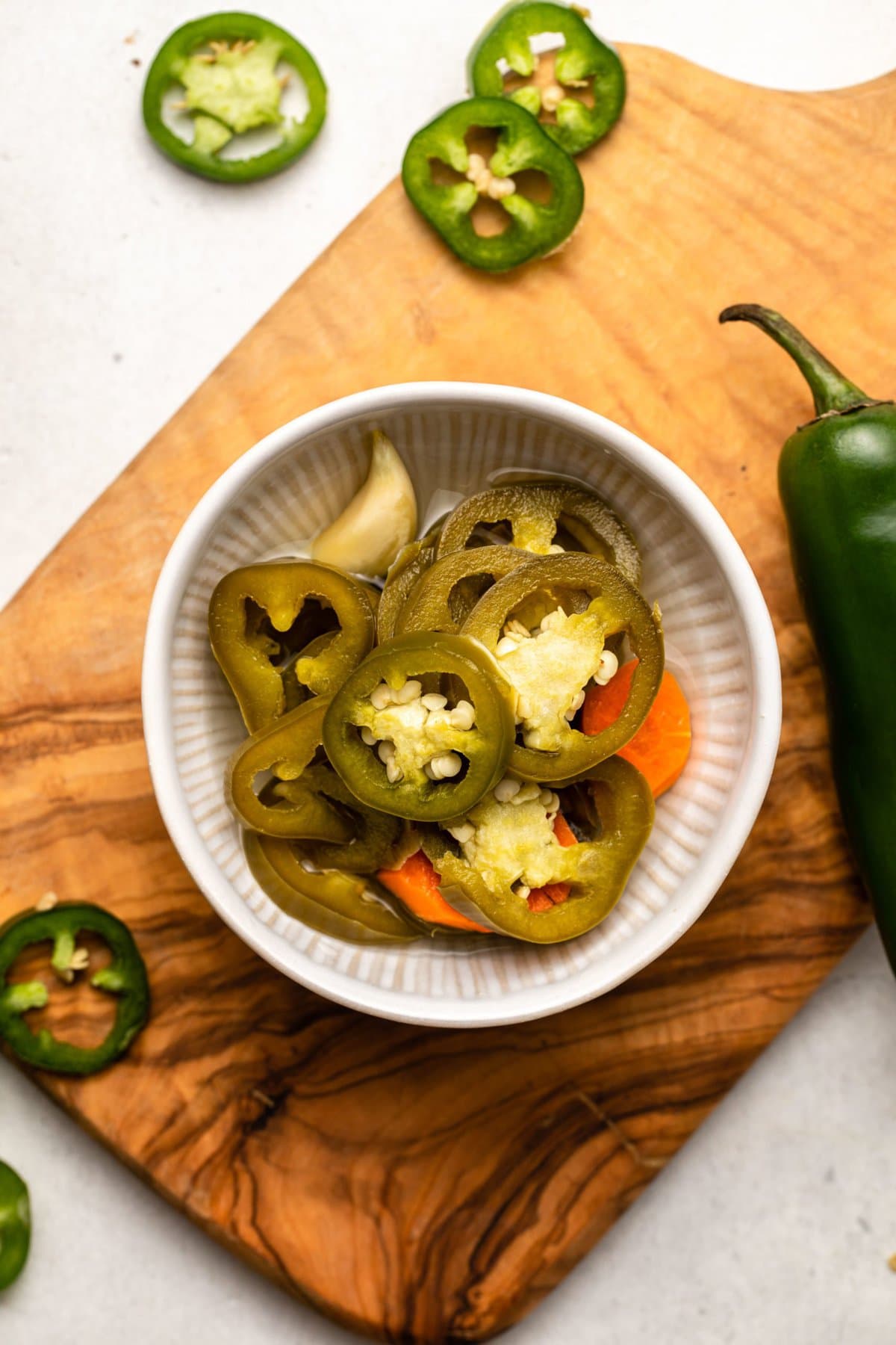 Pickled jalapeños with carrots and garlic in a small white bowl, on a wood cutting board