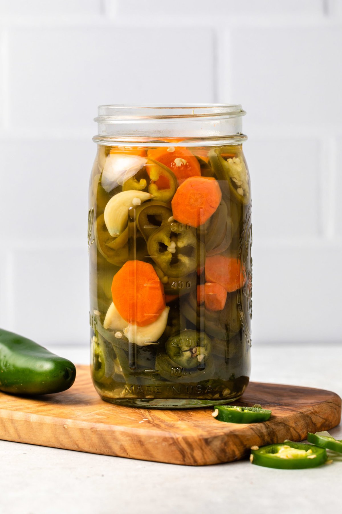 Large glass jar of pickled jalapeños, carrots, and garlic in brine on small wood cutting board. Slices of fresh jalapeños are around the jar.