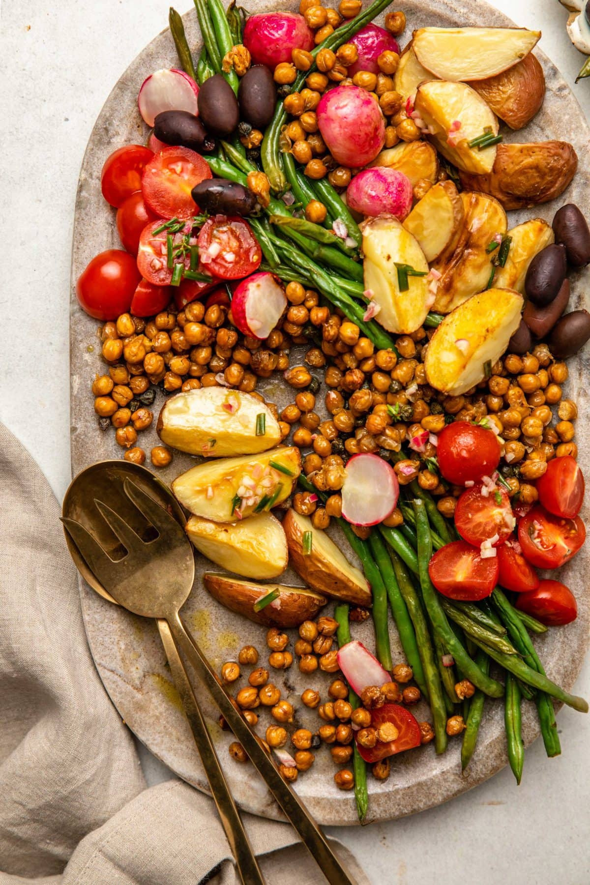 gold serveware on large stone tray. Roasted potatoes, green beans, radishes, tomatoes, olives, crispy chickpeas, and a dressing are on the stone tray