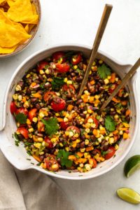 Large white mixing bowl of black bean, corn, tomato, onion, pepper, and cilantro salad with bowl of chips off to the side