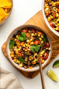 white bowl of tex mex black bean salad topped with fresh cilantro. A gold fork is in the bowl, and the bowl rests on a wood cutting board. Lime wedges and a bowl of tortilla chips are off to the side.