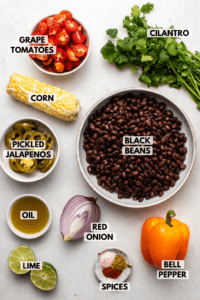 Ingredients for black bean tex mex salad arranged on white background. Clockwise text labels read cilantro, black beans, bell pepper, red onion, spices, lime, oil, pickled jalapeños, corn, and tomatoes