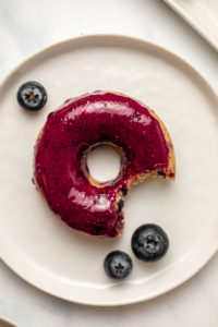close-up photo of glazed blueberry donut on small white plate with bite taken out of it