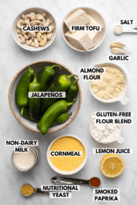 Ingredients for vegan jalapeño poppers arranged in small white bowls on marble background. Clockwise text labels read firm tofu, salt, garlic, almond flour, gluten-free flour blend, lemon juice, smoked paprika, cornmeal, nutritional yeast, non-dairy milk, jalapeños, and cashews