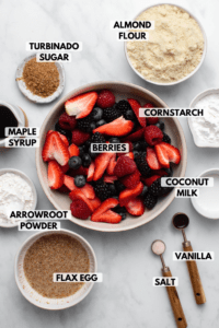 Image of galette ingredients in small white bowls on marble background. Clockwise text labels read almond flour, cornstarch, berries, coconut milk, vanilla, salt, flax egg, arrowroot powder, maple syrup, and turbinado sugar
