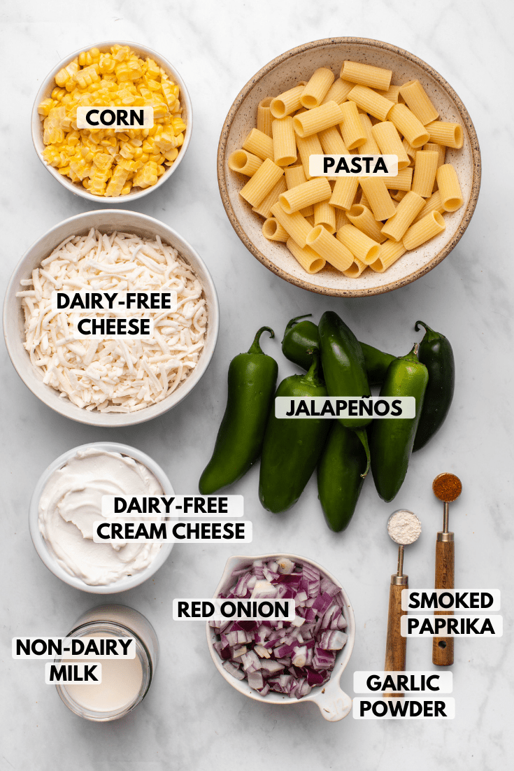 Ingredients for jalapeño popper casserole in small white bowls on marble background. Clockwise text labels read pasta, jalapeños, smoked paprika, garlic powder, red onion, dairy-free milk, dairy-free cream cheese, dairy-free cheese, and corn