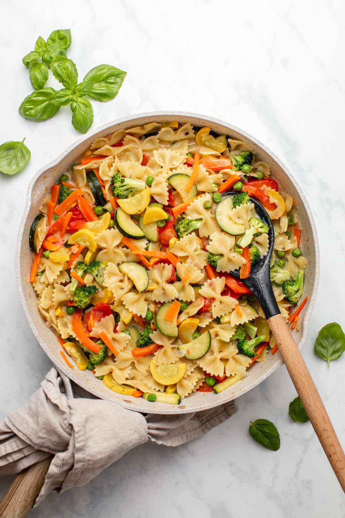 Large sauté pan of cooked pasta primavera with colorful vegetables, with fresh basil on the side