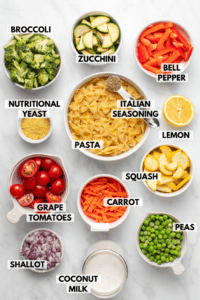 Ingredients for pasta primavera in white bowls on stone background. Clockwise text labels read zucchini, bell pepper, italian seasoning, lemon, squash, carrot, peas, coconut milk, shallot, grape tomatoes, pasta, nutritional yeast, and broccoli