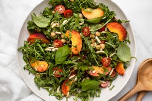Peach, Arugula, & Chickpea Salad topped with grape tomatoes, basil, and almonds in a large white bowl with salad serving spoons off to the side