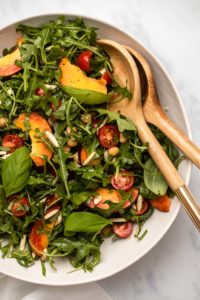 Overhead close-up photo of peach and chickpea salad with arugula, basil, and slivered almonds with wooden salad spoons