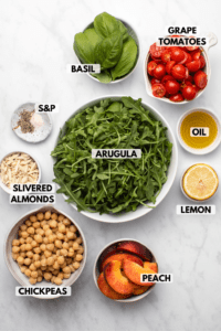 Ingredients for summer peach & chickpea salad in small white bowls on marble background. Clockwise text labels read basil, grape tomatoes, oil, lemon, arugula, peach, chickpeas, slivered almonds, and salt & pepper
