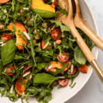 Overhead close-up photo of peach and chickpea salad with arugula, basil, and slivered almonds with wooden salad spoons