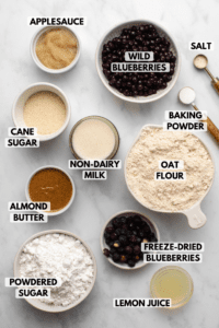 Ingredients for baked blueberry donuts in small white bowls on marble background. Clockwise text labels read wild blueberries, salt, baking powder, oat flour, freeze-dried blueberries, lemon juice, powdered sugar, almond butter, non-dairy milk, cane sugar, and applesauce