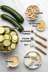 Ingredients for zucchini gratin arranged on marble countertop. Clockwise text labels read cashews, nutritional yeast, smoked paprika, italian seasoning, salt, onion, garlic, cornmeal, and zucchini