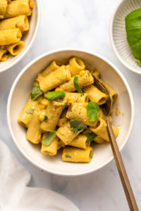 Charred Corn and Basil pasta in small white bowl with a gold fork on a white marble background