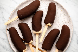 Seven chocolate covered banana pops arranged on a white speckled plate
