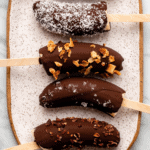 Chocolate covered banana pops decorated with chopped peanuts, coconut, granola, salt, and cacao nibs