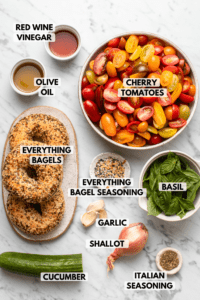 Ingredients for Everything Bagel Panzanella Salad in small white bowls on marble background. Clockwise text labels read cherry tomatoes, everything bagel seasoning, basil, garlic, shallot, italian seasoning, cucumber, everything bagels, olive oil, red wine vinegar