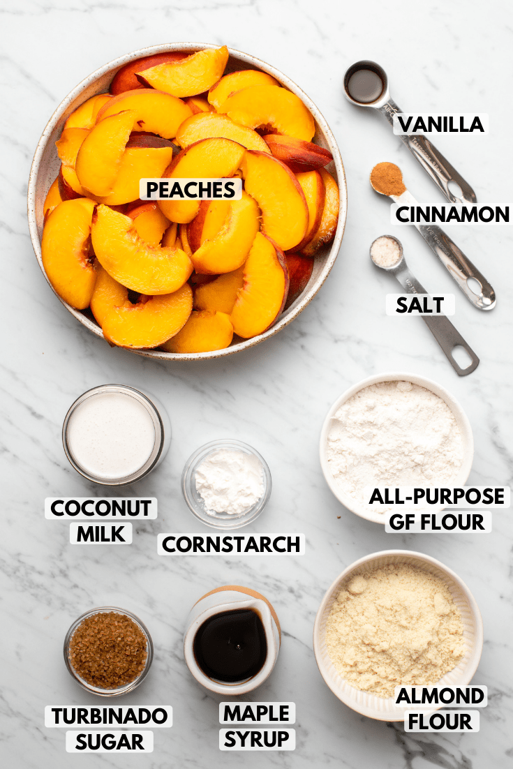 Ingredients for peach cobbler in small white dished on marble background. Clockwise text labels read Vanilla, Cinnamon, Salt, All-Purpose GF Flour, almond flour, maple syrup, turbinado sugar, coconut milk, cornstarch, and peaches