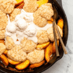 Grilled peach cobbler in cast iron pan with two gold spoons and melting vanilla ice cream