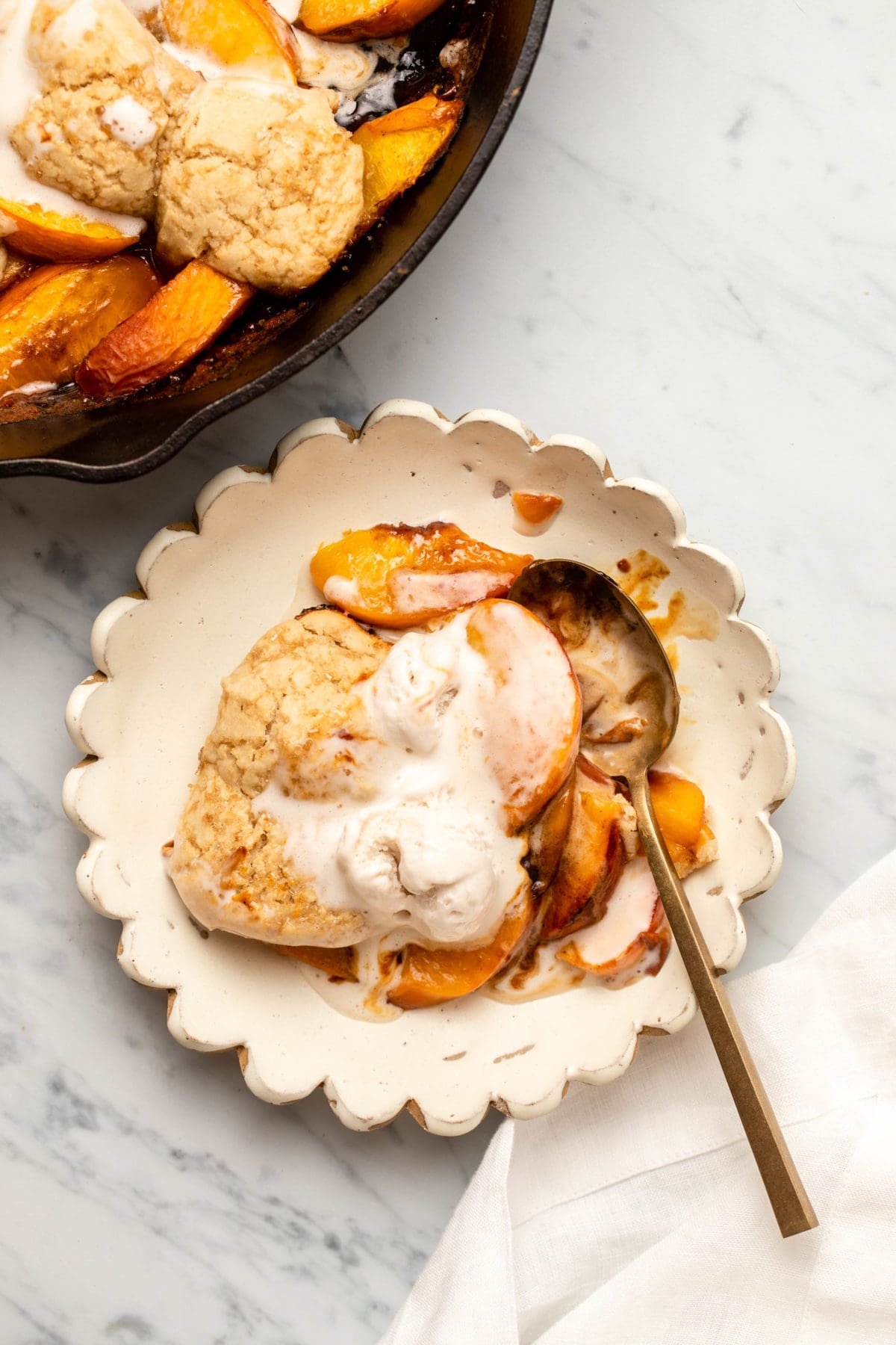 Grilled Peach Cobbler on white scalloped plate with a gold spoon and melted vanilla ice cream on top