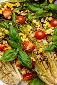 Overhead photo of grilled romaine salad arranged on a large plate with charred corn, tomatoes, basil, and toasted pine nuts