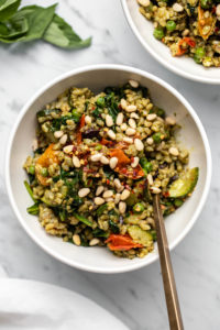 Close up photo of pesto fried rice with colorful cooked tomatoes in a small white bowl topped with pine nuts and a gold fork