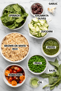 Ingredients for pesto fried rice in small white bowls on a marble countertop. Clockwise ingredient labels read garlic, zucchini, pesto, peas, basil, oil, tomatoes, brown or white rice, spinach, and kalamata olives