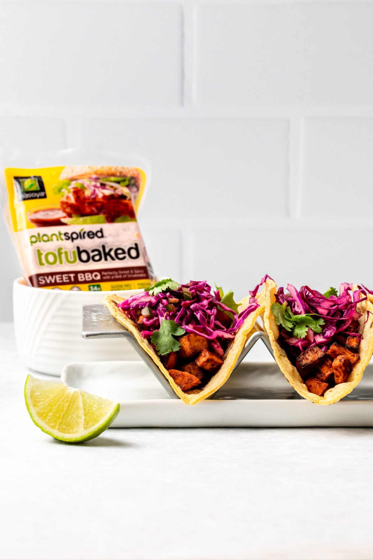 Two tacos topped with red cabbage slaw with a package of Nasoya's Plantspired Sweet BBQ TofuBaked in the background