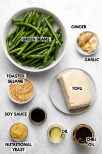 Ingredients for green bean stir fry in small bowls on marble background. Clockwise text labels read ginger, garlic, tofu, chili oil, oil, nutritional yeast, soy sauce, toasted sesame, and green beans