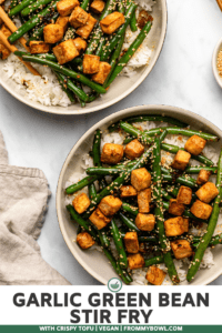 Two large bowls of garlic green bean stir fry with crispy tofu over white rice. A small bowl of toasted sesame seed sits off to the side with a pair of wooden chopsticks
