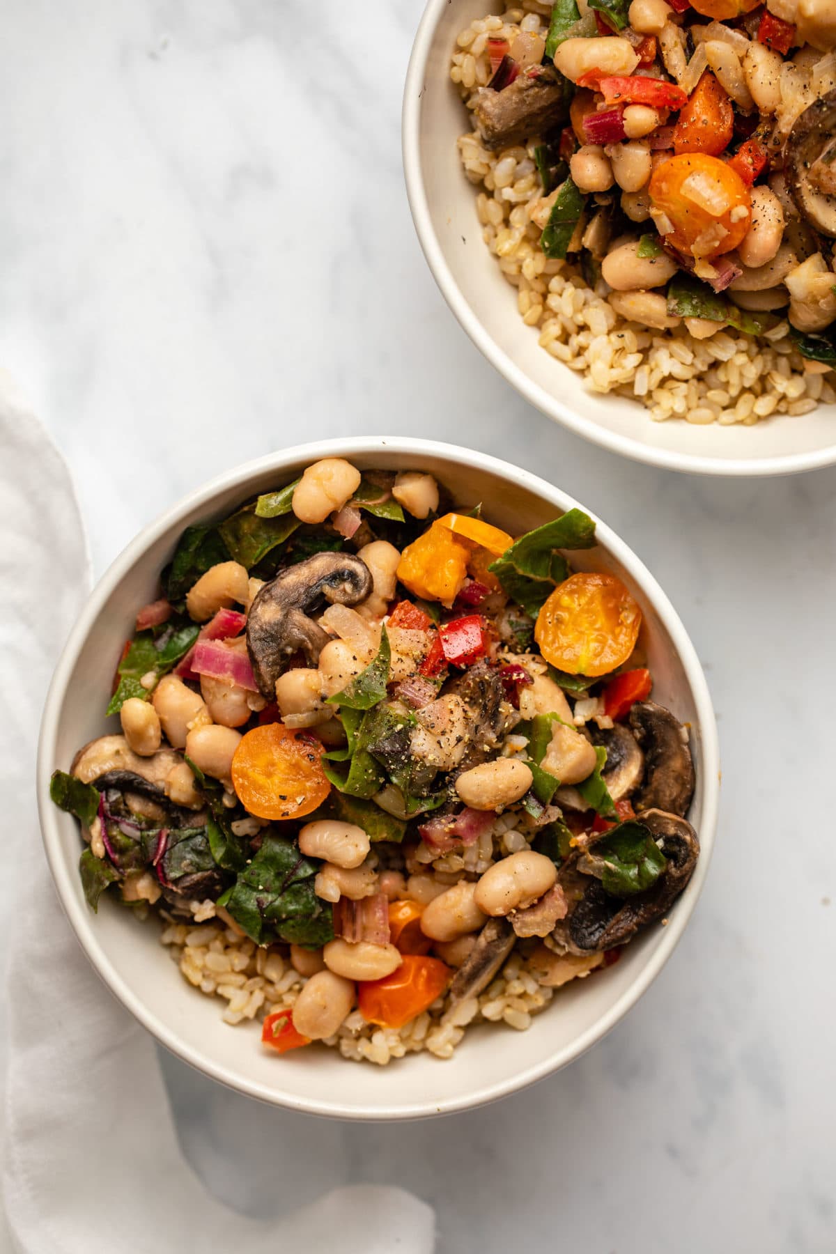 Two white bowls of white bean stir fry with brown rice on marble countertop