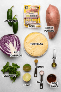 Ingredients for tacos arranged on marble background. Clockwise text labels read sweet potato, tortillas, oil, bbq sauce, salt, cumin, lime, cilantro, red cabbage, jalapeno, tofu