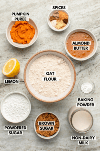 Ingredients for pumpkin chai spiced donuts in small bowls on stone background. Clockwise text labels read spices, almond butter, oat flour, baking powder, non-dairy milk, brown sugar, powdered sugar, lemon, and pumpkin puree