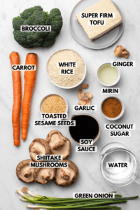 Ingredients for teriyaki tofu rice bake in small white bowls on marble background. Clockwise text labels read super firm tofu, ginger, mirin, garlic, coconut sugar, soy sauce, water, green onion, shiitake mushrooms, toasted sesame seeds, white rice, carrot, and broccoli