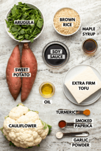Ingredients for nourish bowls with maple turmeric tofu in small bowls on stone background. Clockwise text labels read brown rice, maple syrup, extra firm tofu, turmeric, smoked paprika, garlic powder, cauliflower, oil, sweet potato, arugula, and soy sauce