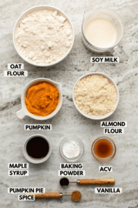 Ingredients for pumpkin spice waffles in small white bowls on stone background. Clockwise text labels read soy milk, almond flour, acv, vanilla, pumpkin pie spice, baking powder, maple syrup, pumpkin, and oat flour