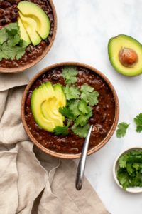 Two bowls of chipotle black bean chili topped with avocado and cilantro on white marble background