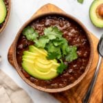 Bowl of chipotle black bean chili topped with avocado and cilantro on wood cutting board