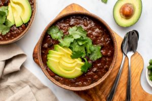 Bowl of chipotle black bean chili topped with avocado and cilantro on wood cutting board
