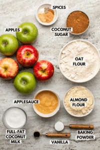 Ingredients for vegan apple cake in small white bowls on stone background. Clockwise text labels read spices, coconut sugar, oat flour, almond flour, baking powder, vanilla, applesauce, full-fat coconut milk, and apples