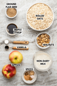 Ingredients for baked apple cinnamon oatmeal in small white bowls on stone countertop. Clockwise text labels read rolled oats, walnuts, non-dairy milk, spices, apples, vanilla, maple syrup, and ground flax seed