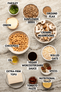Ingredients for vegan meatballs in small white bowls on stone background. Clockwise text labels read ground flax, cremini mushroooms, rolled oats, spices, vegan worcestershire sauce, nutritional yeast, tomato paste, extra-firm tofu, oil, chickpeas, onion, parsley, and sunflower seeds